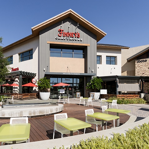 The exterior of Finney's Crafthouse shows a modern building with a peaked roof. The patio features lime-green chairs and white tables under a clear blue sky.